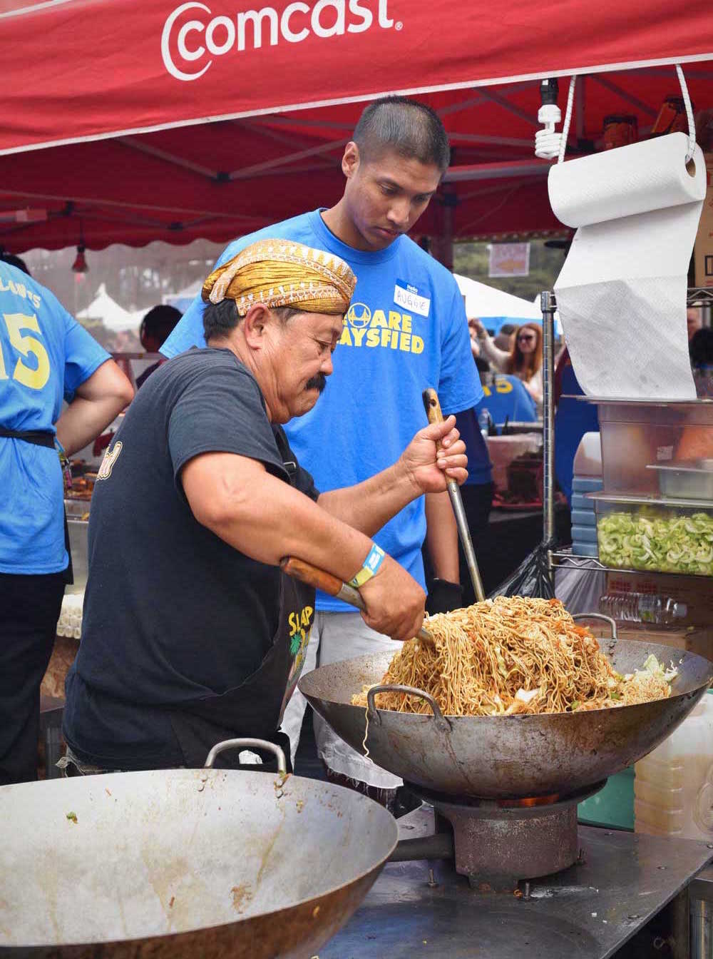 Feldo Nartapura's father, a veteran cook, himself, teaches a Sataysfied volunteer how to mix a giant wok-full of noodles.